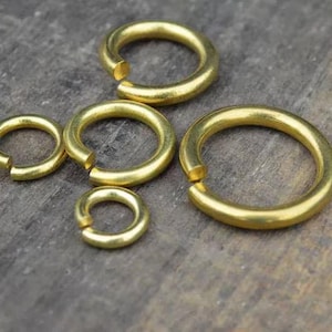 Può includere: Cinque anellini di giunzione in metallo color oro di varie dimensioni, ognuno caratterizzato da un singolo taglio aperto lungo la fascia circolare. Gli anelli hanno una finitura liscia e lucida e sono disposti in gruppo, mostrando diametri.