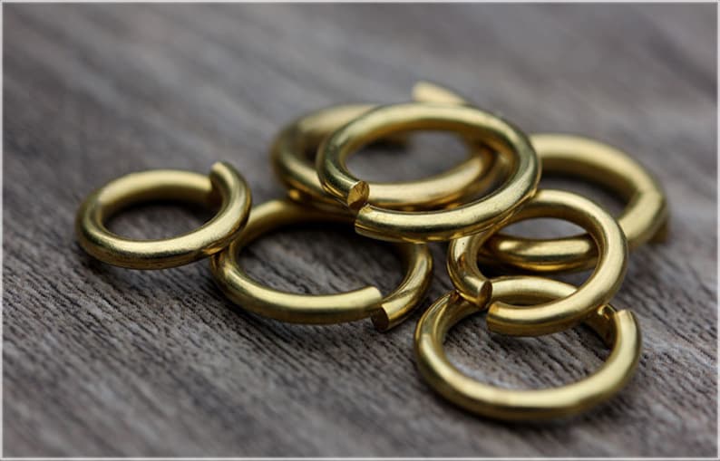 May include: A group of six gold-colored metal jump rings, all of the same size, are arranged in a pile on a wooden surface.