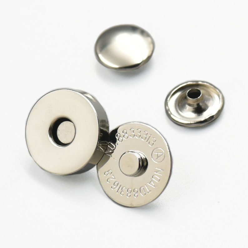 May include: A collection of silver magnetic snap fasteners. The image shows two circular pieces with a central hole, and two smaller, rounded pieces. The larger pieces are marked with the numbers "NO.8833313" and "8831628".