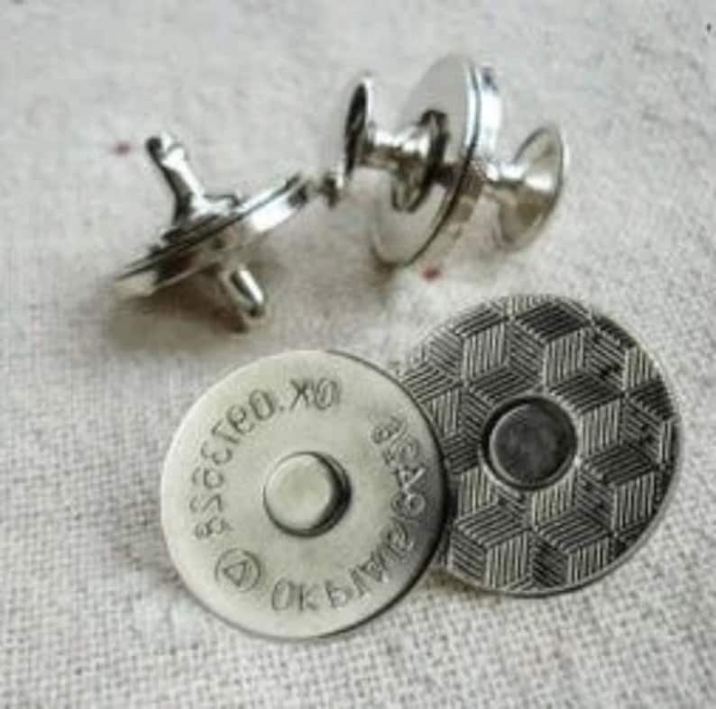 May include: Silver magnetic snap fasteners. One is open, showing the two parts. The other is closed. Two circular pieces have text and a geometric pattern. The snaps are on a light-coloured fabric.