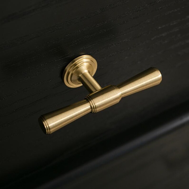 Op de afbeelding: Een goudkleurige T-vormige kastknop met een ronde basis. De knop is gemaakt van metaal en heeft een gladde, gepolijste afwerking.