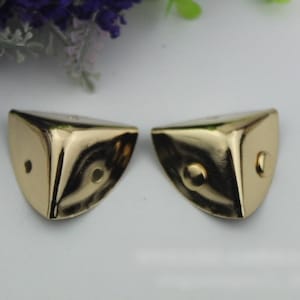 May include: Two gold-colored metal corner protectors with a rounded edge and a small raised dot in the center of each.