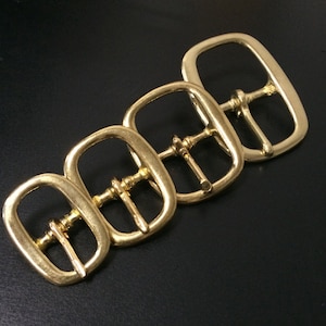 May include: Four brass-colored belt buckles arranged diagonally on a black surface. Each buckle has a rectangular frame with a central bar and a pin for securing a belt. The buckles vary slightly in size, with the largest at the top right.
