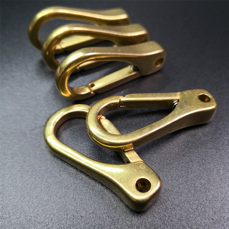 May include: Four brass-colored carabiner clips are arranged on a dark surface. The clips have a spring-loaded gate and a rounded shape. The metal has a polished finish, reflecting light. The carabiners are likely used for securing items.