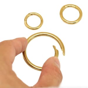May include: Gold-colored metal spring rings in various sizes. One large ring is open, held by a hand, while two smaller rings are closed. The rings are on a white background.