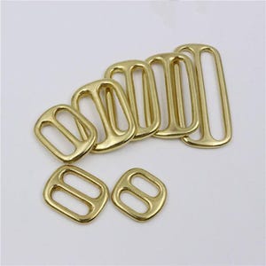 May include: A collection of eight gold-toned metal strap adjusters of varying sizes. Each piece features a rectangular frame with a central bar, designed for sliding fabric straps through to adjust length. The finish is smooth and metallic.