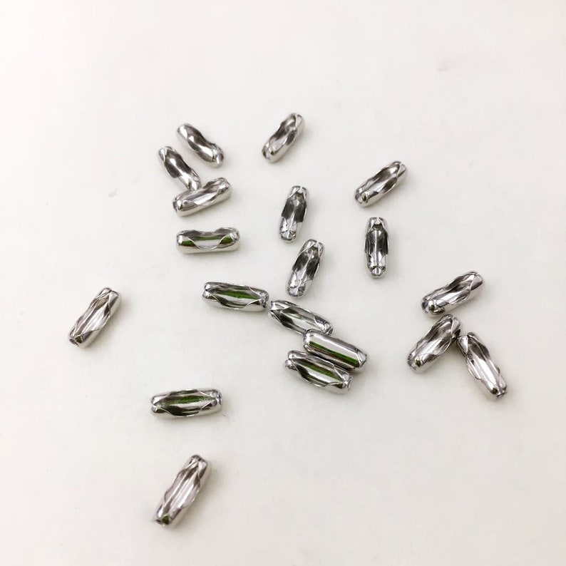 May include: A scattering of silver-toned metal chain connectors, also known as chain links, on a white surface. These connectors are used to join two pieces of chain together.