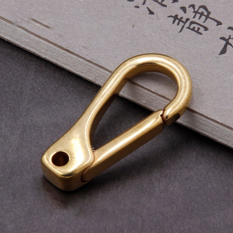 May include: A solid brass carabiner clip with a spring-loaded gate. The clip has a rectangular base with a drilled hole and a curved top. The background is a gray surface with some text.