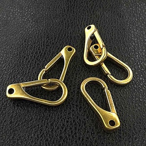 May include: Four brass-colored, spring-loaded snap hooks. Each hook has a rounded, teardrop-shaped body with a small hole at the end and a spring-loaded clasp. The hooks are arranged on a dark, textured surface.