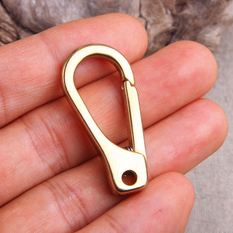 May include: A gold-colored, spring-loaded carabiner clip. The clip has a curved top and a straight, rectangular body with a small hole at the bottom. The clip is designed to attach to a variety of items.