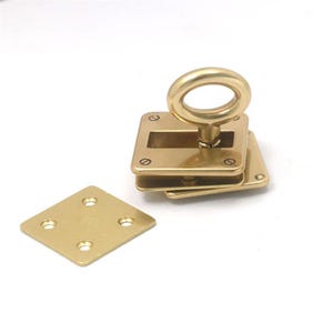 Könnte beinhalten: Ein messingfarbenes, quadratisches Beschlagteil mit einem Ringgriff. Die Hardware besteht aus mehreren Schichten und enthält eine separate rautenförmige Platte mit vier Löchern. Die Hardware ist wahrscheinlich für Möbel oder eine ähnliche Anwendung.