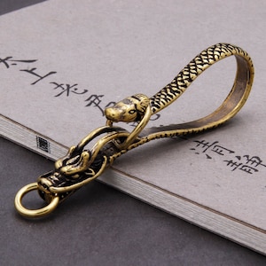 May include: A gold-toned metal key chain with a dragon head and a snake body. The snake's head is at the top of the loop and the dragon's head is at the bottom.