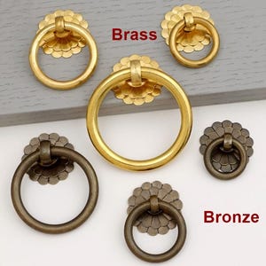 Könnte beinhalten: Mehrere Messing- und Bronzeringgriffe mit floralen Rückplatten werden gezeigt. Das Wort "Brass" steht in Rot über den goldfarbenen Griffen, und "Bronze" steht in Rot über den dunkleren Griffen. Diese Griffe sind wahrscheinlich für Schränke oder Schubladen.