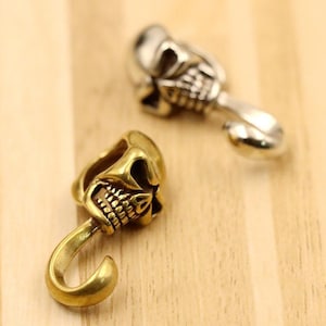 May include: Two metal skull-shaped beads, one in a polished silver tone and one in a brushed gold tone. Each bead features a detailed skull design with a curved hook or loop extending from the base, crafted from solid metal with visible textures.