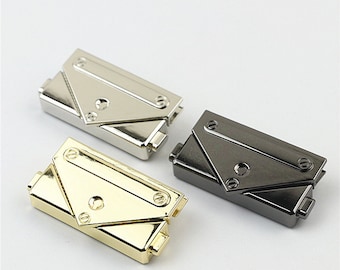 May include: Three metal purse clasps in silver, gold, and gunmetal gray. Each clasp has a rectangular base with a hinged, triangular flap secured by screws. The clasps are designed for bags and purses.