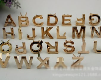 May include: Gold-colored metal alphabet letter charms with a pointed back for attaching to a project. The letters are arranged in alphabetical order.