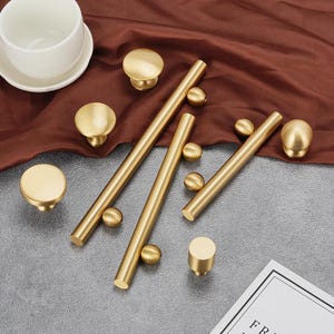 May include: A set of gold-toned cabinet knobs and drawer pulls. The knobs are round and have a smooth finish. The pulls are long and have a cylindrical shape with a decorative ball at each end.