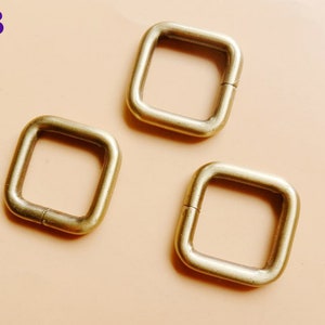 May include: Three gold-colored metal square rings with a rounded edge. The rings are open on one side and can be used for attaching items.