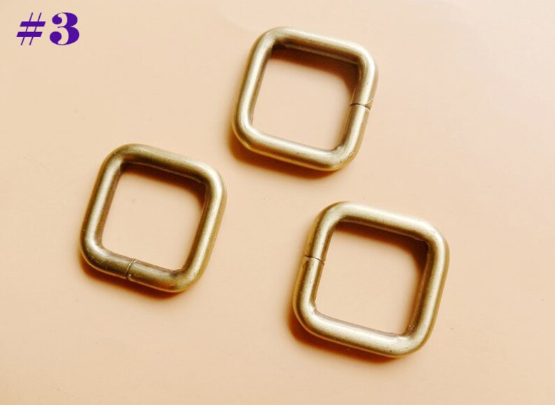 May include: Three gold-colored metal square rings with a rounded edge. The rings are open on one side and can be used for attaching items.