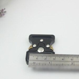 May include: Black plastic buckle with gold accents, measured with a ruler. The buckle is approximately 1 1/2 inches wide.
