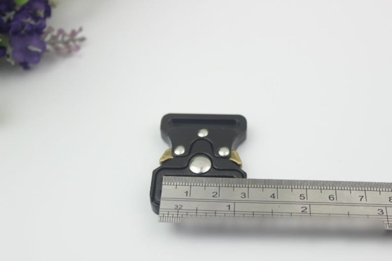 May include: Black plastic buckle with gold accents, measured with a ruler. The buckle is approximately 1 1/2 inches wide.