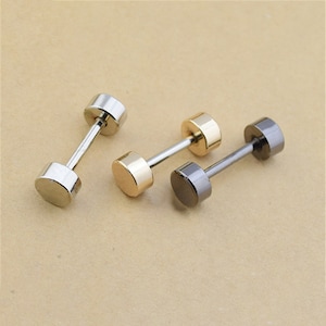 May include: Three dumbbell-shaped stud earrings in silver, gold, and gunmetal colors. Each earring features a cylindrical barbell connecting two circular end pieces. The earrings are displayed on a neutral background.