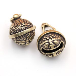 May include: Two antique brass bells with detailed embossed patterns. One bell is rounded with a loop, the other spherical. The bells have a warm, aged finish, suggesting a vintage or handcrafted origin. The intricate designs are visible on both.