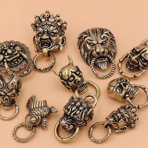 May include: A collection of ten brass-toned metal door knockers and decorative pulls featuring various sculpted designs including lion heads, horned demon masks, skulls, and a fish, each with an attached circular ring for pulling or hanging.