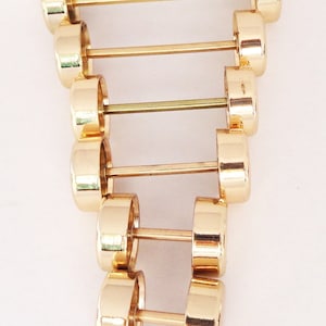 May include: A collection of gold-colored barbell earrings, each with a cylindrical weight on either end connected by a straight bar. The earrings are arranged in a vertical line against a white background, showcasing their reflective, polished surface.