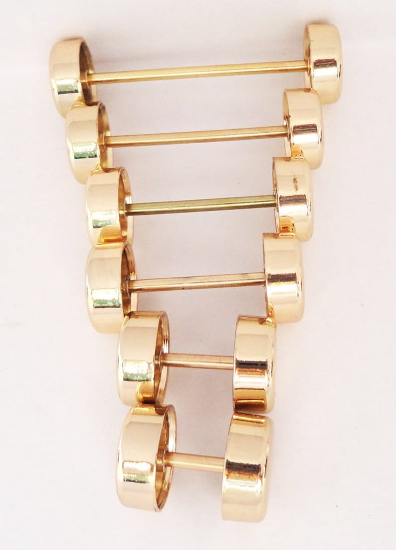 May include: A collection of gold-colored barbell earrings, each with a cylindrical weight on either end connected by a straight bar. The earrings are arranged in a vertical line against a white background, showcasing their reflective, polished surface.