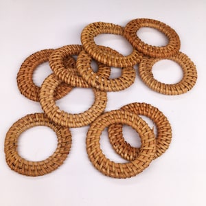 May include: A collection of woven rattan napkin rings. The rings are circular, with a light brown color and a textured surface. The napkin rings are arranged on a white background.