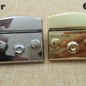 Könnte beinhalten: Zwei Metallverschlüsse, einer silberfarben und einer goldfarben, mit jeweils drei runden Nieten. Die Verschlüsse sind geschlossen und haben eine rechteckige Form.