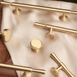 Op de afbeelding: Een set van vijf gouden kastbeslag trekt en knoppen. De trekt zijn lang en slank met een afgeronde uiteinde. De knoppen zijn rond met een verhoogde rand. De hardware is gerangschikt op een licht beige oppervlak.