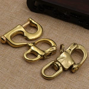 May include: Three polished brass swivel snap hooks of varying designs. The left hook features a screw-lock gate mechanism, while the center and right hooks are spring-loaded swivel clips with integrated loops for attachment to straps or cords.