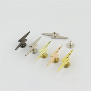 May include: Five lightning bolt shaped metal studs in different finishes: black, silver, gold, rose gold, and brass. Each stud has a screw-on back.