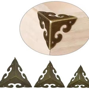 Peut inclure: Supports d'angle décoratifs de couleur bronze antique. Les supports triangulaires présentent des motifs de volutes. L'image montre un gros plan d'un support fixé sur du bois, ainsi que trois autres supports.
