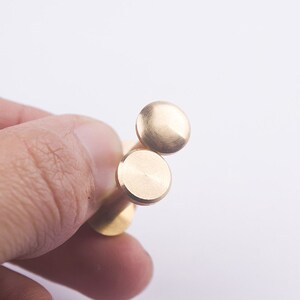 May include: Two gold-coloured metal rivets with a round head. The rivets are held in a hand.