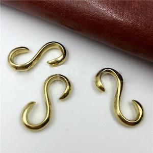 May include: Three brass-colored S-hooks are displayed against a white surface, with a brown leather background. The hooks are curved and have a smooth, polished finish. They are likely used for hanging or connecting items.