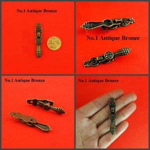 May include: An antique bronze metal drop pull handle featuring an elongated, ornate design with a central circular floral motif and a tapered bottom. The hardware is labeled No.1 Antique Bronze and is shown next to a 5 coin for scale.