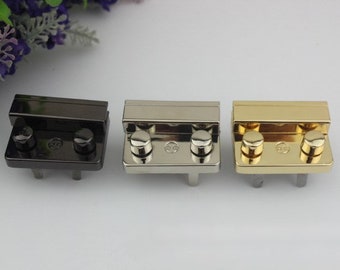 May include: Three metal purse clasps in black, silver, and gold. Each clasp has a rectangular body with two rounded knobs and two small posts. The clasps are arranged horizontally against a white background.