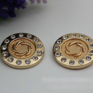 May include: Two gold-toned round buttons with a swirl design in the center and a circle of clear rhinestones around the edge.