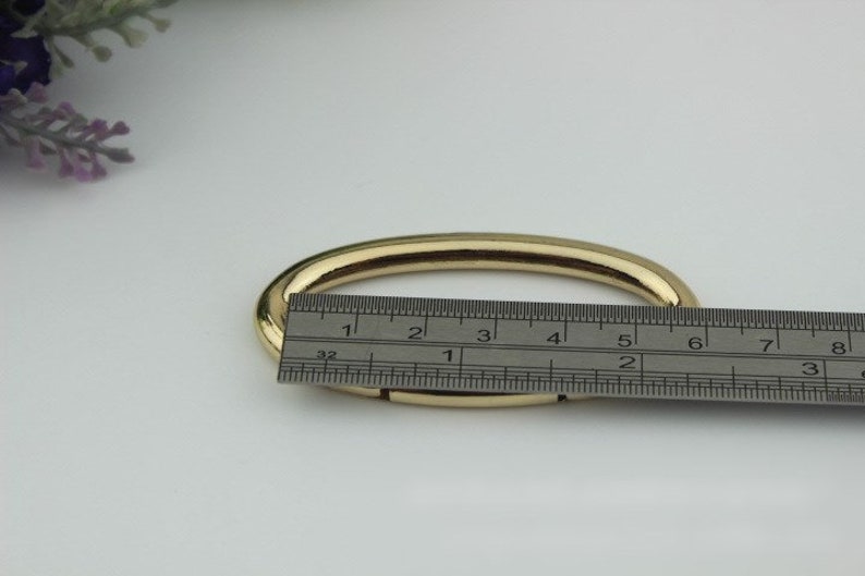 May include: A gold-colored metal oval ring, measuring approximately 3 inches in diameter, is shown on a white surface with a ruler for scale.