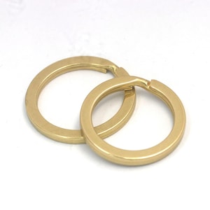 May include: Two gold-colored key rings are interlocked, set against a white background. The rings are circular with a flat profile and a slight sheen, suggesting a metallic finish. The key rings are designed to hold keys or other small items.