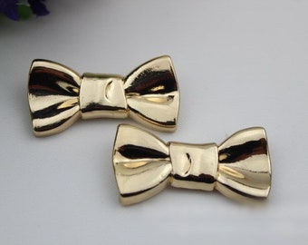 May include: Two gold-colored bow-shaped metal embellishments. The bows are about 2 inches long and 1 inch wide.