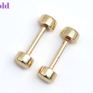 May include: Two gold-colored barbell stud earrings. Each earring has a cylindrical post connecting two rounded, flat ends. The word "Gold" is in the upper left corner in a purple font. The earrings are on a white background.