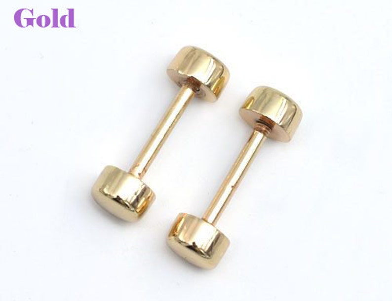 May include: Two gold-colored barbell stud earrings. Each earring has a cylindrical post connecting two rounded, flat ends. The word "Gold" is in the upper left corner in a purple font. The earrings are on a white background.