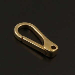 May include: A solid brass carabiner clip with a spring-loaded gate. The clip has a smooth, polished finish and a rounded top. The overall shape is a classic oval with a rectangular base and a small hole for attachment.