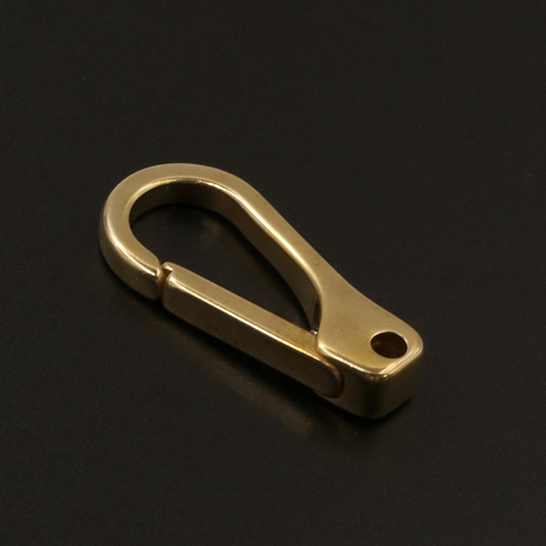 May include: A solid brass carabiner clip with a spring-loaded gate. The clip has a smooth, polished finish and a rounded top. The overall shape is a classic oval with a rectangular base and a small hole for attachment.