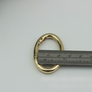 May include: A gold-colored metal oval-shaped ring with a spring-loaded clasp. The ring is measured with a ruler, showing it is approximately 1 inch in diameter.