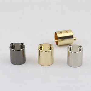 May include: A collection of metallic cylindrical hardware components in various finishes: gunmetal, gold, and silver. Each cylinder has two small screws on top, with one gold cylinder having a hollow, open design.